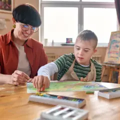 Lehrerin unterstützt Kind mit Down-Syndrom beim Malen. Ein Beispiel für Förderung bei Autismus bei Kleinkindern.