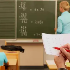 Lehrerin erklärt Mathe an Tafel. Ein Blatt mit Namen und Kästchen liegt bereit, wie bei dienstliche Beurteilung Lehrer Beispiele.
