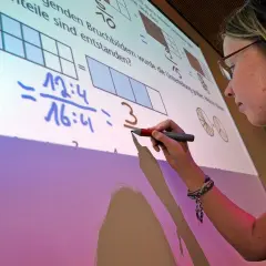 Ein Mädchen schreibt auf einem interaktiven Whiteboard, das Matheaufgaben zeigt. Solche Whiteboards sind moderne Tafeln für den Unterricht.