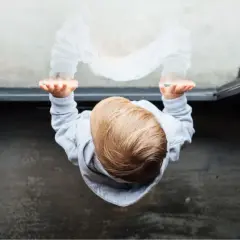 Kind weint nach einem Jahr im Kindergarten. Die Hände des Kindes sind gegen eine Scheibe gedrückt, sein Spiegelbild schaut traurig zurück.