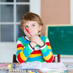 Ein Junge mit einem roten Stift im Mund denkt nach. Seine Kleidung und die Lernmaterialien deuten auf eine mögliche **Hochbegabung bei Kindern** hin.