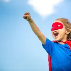 Ein kleines Mädchen im Superheldenkostüm mit rotem Cape und Maske, bereit für den **ersten Schultag**.