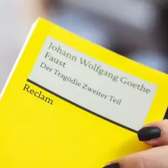 Ein gelbes Buch von Reclam mit Goethes Faust, Teil 2. Eine Hand mit schwarzen Nägeln hält es. Textanalyse Deutsch.