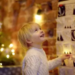 Kleiner Junge freut sich über Adventskalender mit Grundschule Ideen. Weihnachtsbaum und Lichter im Hintergrund.