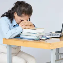 Erschöpfte Frau am Schreibtisch, fragt sich: "Soll ich Lehrer werden?" Bücher, Laptop und Brille liegen bereit.