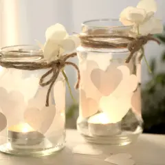 Zwei selbstgemachte Windlichter aus Gläsern, verziert mit Herzen und Blumen, strahlen warmes Kerzenlicht aus. Perfekt zum Windlichter basteln!