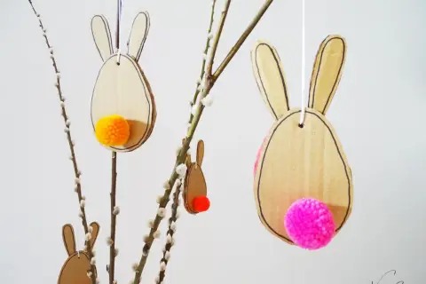 Osterhasen aus Pappe mit bunten Bommeln als Schwänzen, perfekt zum Basteln Ostern Kinder. Hängen an Weidenkätzchen-Zweigen.