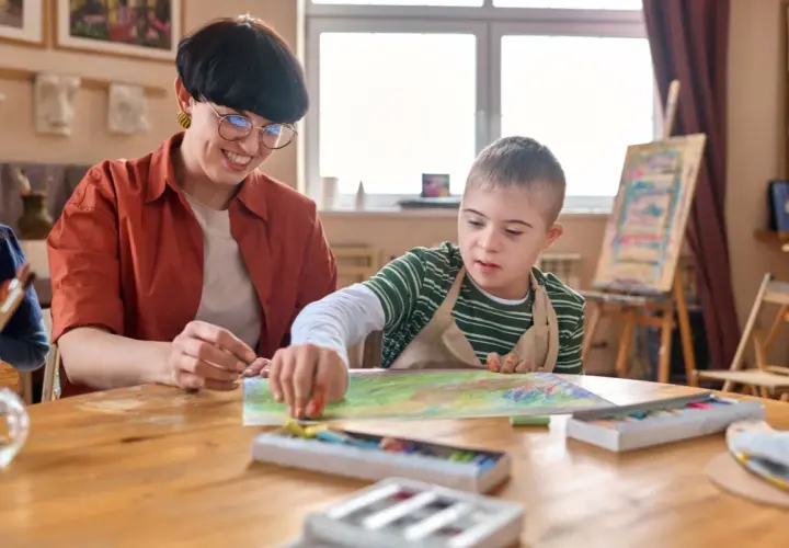 Lehrerin unterstützt Kind mit Down-Syndrom beim Malen. Ein Beispiel für Förderung bei Autismus bei Kleinkindern.