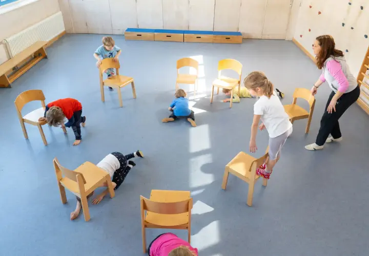 Kinder spielen in der Turnhalle, nutzen Stühle für kreative Ideen und Bewegung.
