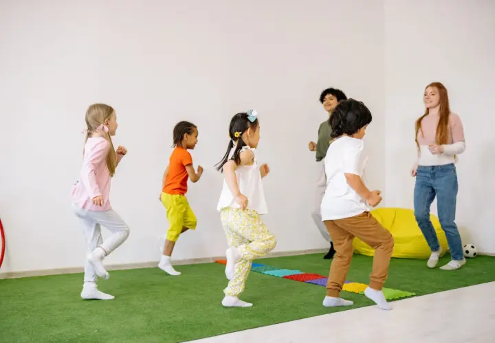 Kindergartenkinder rennen fröhlich über den grünen Teppich, als ob sie Tiere wären. Die Erzieherinnen machen mit bei den lustigen Bewegungsspielen.
