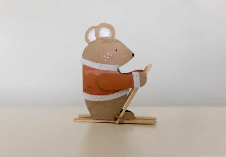 Süße Maus beim Skifahren, ein tolles Beispiel für Basteln mit Papprollen. Sie trägt einen orangen Pullover und hält Skistöcke.