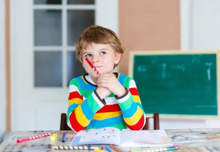 Ein Junge mit einem roten Stift im Mund denkt nach. Seine Kleidung und die Lernmaterialien deuten auf eine mögliche **Hochbegabung bei Kindern** hin.