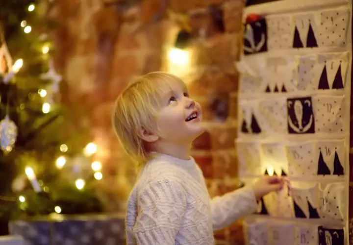 Kleiner Junge freut sich über Adventskalender mit Grundschule Ideen. Weihnachtsbaum und Lichter im Hintergrund.