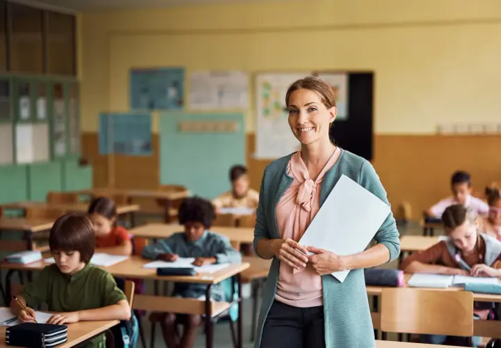 Lehrerberuf pro contra: Eine lächelnde Lehrerin mit Unterlagen steht vor einer Klasse voller konzentrierter Schüler.