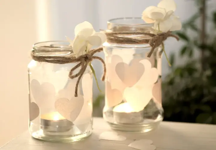 Zwei selbstgemachte Windlichter aus Gläsern, verziert mit Herzen und Blumen, strahlen warmes Kerzenlicht aus. Perfekt zum Windlichter basteln!