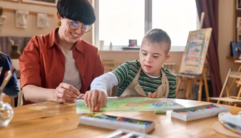 Lehrerin unterstützt Kind mit Down-Syndrom beim Malen. Ein Beispiel für Förderung bei Autismus bei Kleinkindern.