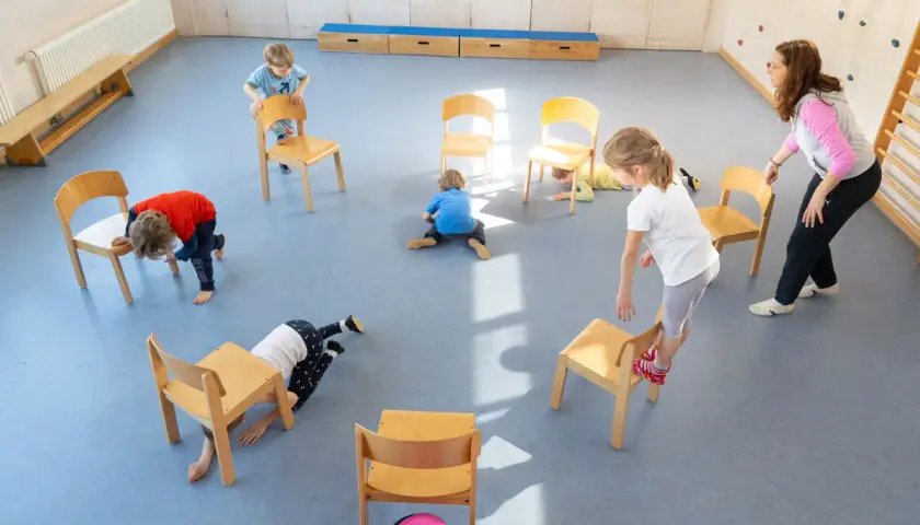 Kinder spielen in der Turnhalle, nutzen Stühle für kreative Ideen und Bewegung.