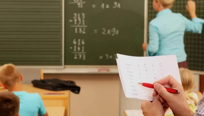 Lehrerin erklärt Mathe an Tafel. Ein Blatt mit Namen und Kästchen liegt bereit, wie bei dienstliche Beurteilung Lehrer Beispiele.