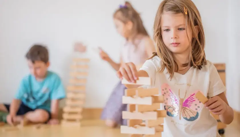 Kinder gestalten eine Bauecke im Kindergarten. Ein Mädchen baut konzentriert einen Turm aus Holzklötzen.