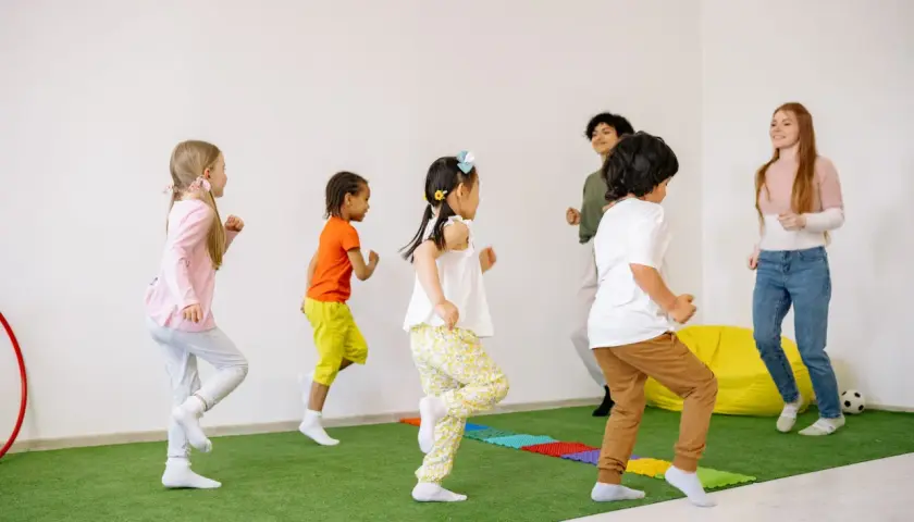 Kindergartenkinder rennen fröhlich über den grünen Teppich, als ob sie Tiere wären. Die Erzieherinnen machen mit bei den lustigen Bewegungsspielen.