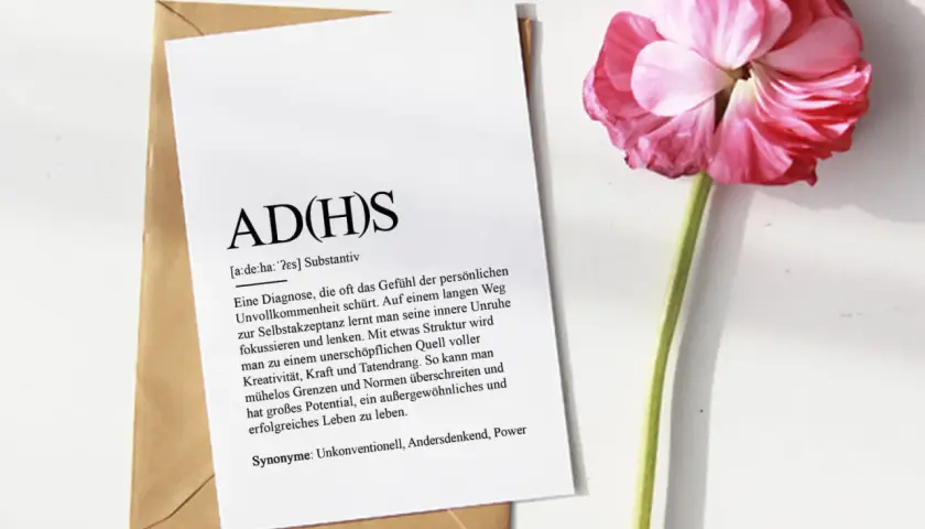 ADHS-Diagnostik online: Ein Blatt mit der Definition von ADHS liegt auf einem Umschlag, daneben eine rosa Blüte.