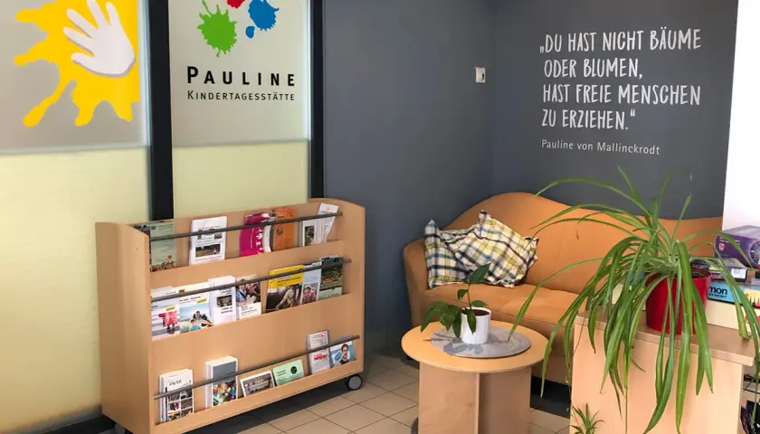 Einladende Ideen für den Eingangsbereich einer Kita: Bücherregal mit Broschüren, gemütliche Sitzecke mit Zitatwand und Pflanzen.