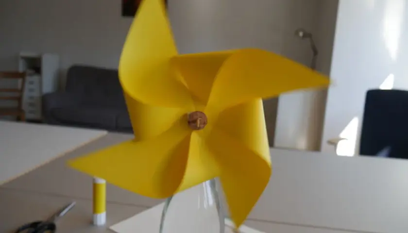 Ein gelbes Windrad, das man leicht selber bauen kann, steht in einer Glasflasche. Schere und Klebestift liegen bereit.