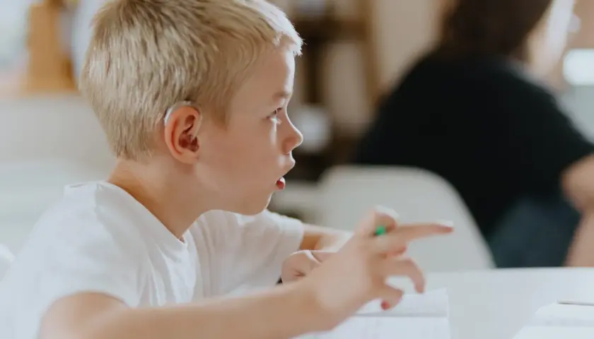 Junge mit Hörgerät, der auf ein Buch zeigt. Mögliche ADHS-Ursachen werden diskutiert, während er sich konzentriert.
