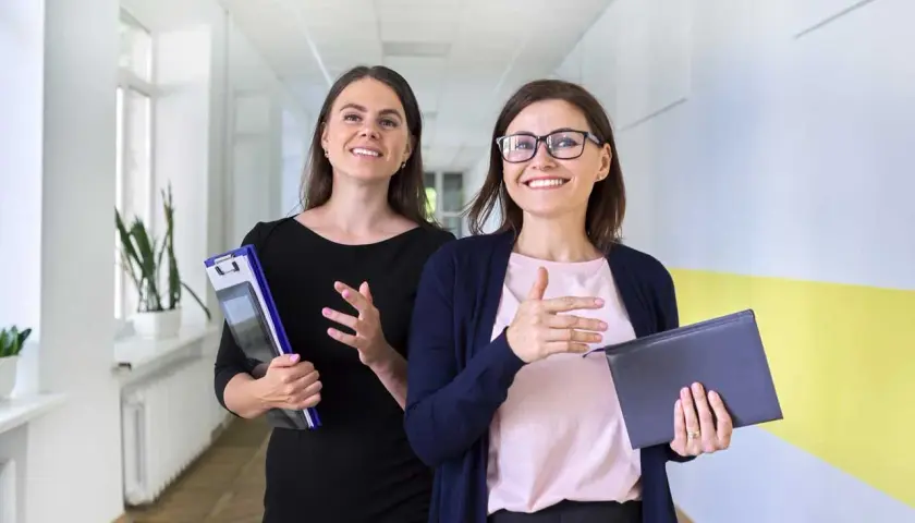 Zwei Frauen gehen lächelnd einen Flur entlang. Die eine hält eine Mappe, die andere ein Buch. Sie könnten eine Lehrerin und eine Referendarin sein, die sich über den Schulalltag austauschen.