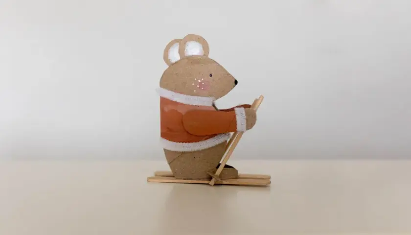 Süße Maus beim Skifahren, ein tolles Beispiel für Basteln mit Papprollen. Sie trägt einen orangen Pullover und hält Skistöcke.