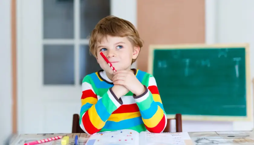 Ein Junge mit einem roten Stift im Mund denkt nach. Seine Kleidung und die Lernmaterialien deuten auf eine mögliche **Hochbegabung bei Kindern** hin.