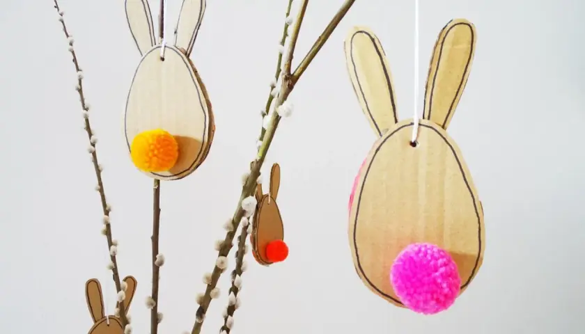 Osterhasen aus Pappe mit bunten Bommeln als Schwänzen, perfekt zum Basteln Ostern Kinder. Hängen an Weidenkätzchen-Zweigen.