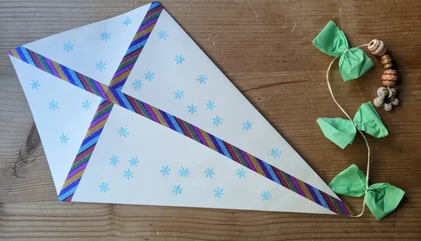 Ein selbstgebastelter Drachen aus Tonpapier mit Schneeflocken und bunten Bändern. Die Schnur hat kleine grüne Schleifen und Holzperlen.