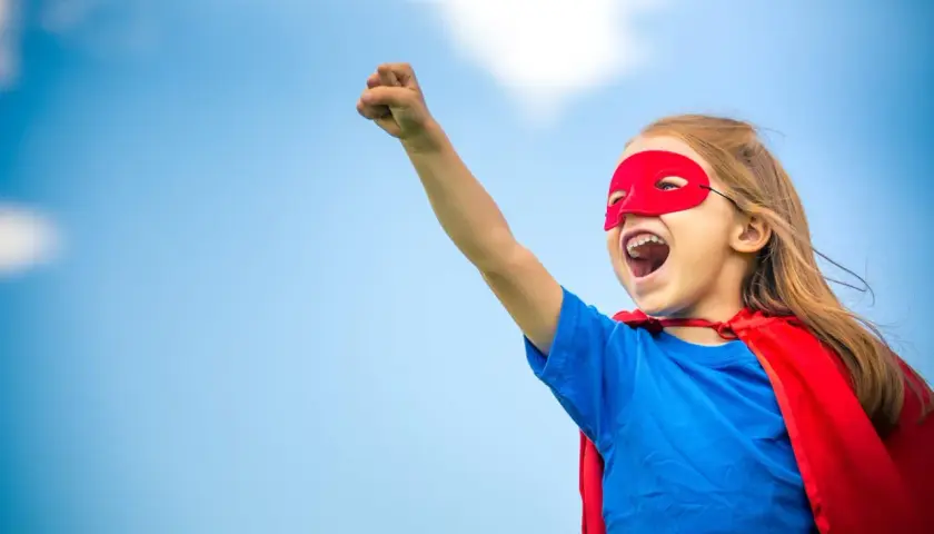 Ein kleines Mädchen im Superheldenkostüm mit rotem Cape und Maske, bereit für den **ersten Schultag**.