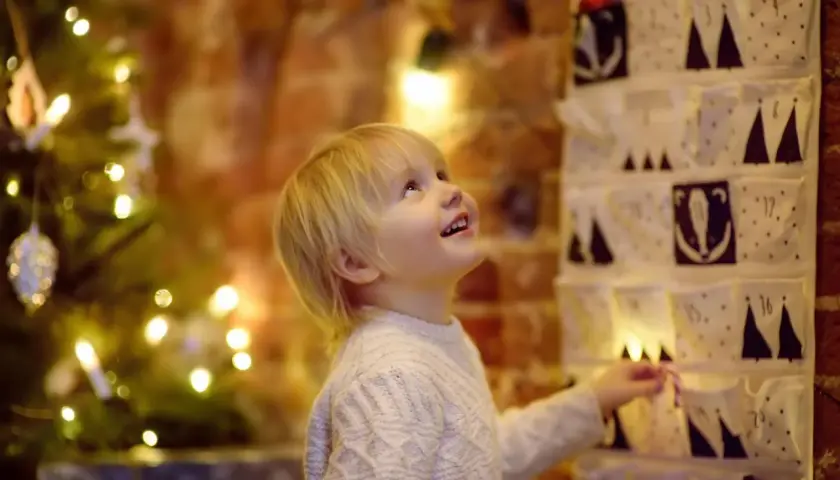 Kleiner Junge freut sich über Adventskalender mit Grundschule Ideen. Weihnachtsbaum und Lichter im Hintergrund.
