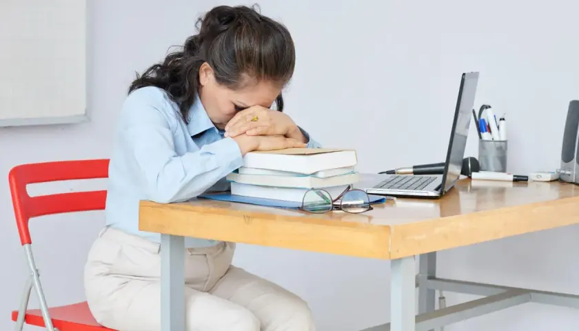 Erschöpfte Frau am Schreibtisch, fragt sich: "Soll ich Lehrer werden?" Bücher, Laptop und Brille liegen bereit.