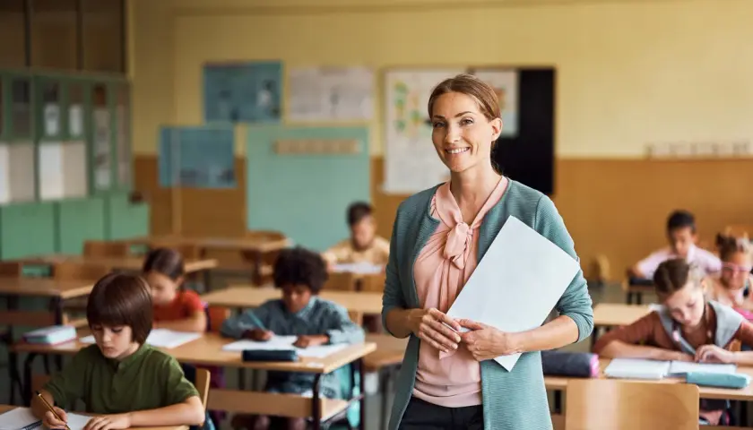 Lehrerberuf pro contra: Eine lächelnde Lehrerin mit Unterlagen steht vor einer Klasse voller konzentrierter Schüler.