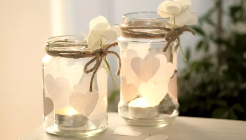 Zwei selbstgemachte Windlichter aus Gläsern, verziert mit Herzen und Blumen, strahlen warmes Kerzenlicht aus. Perfekt zum Windlichter basteln!