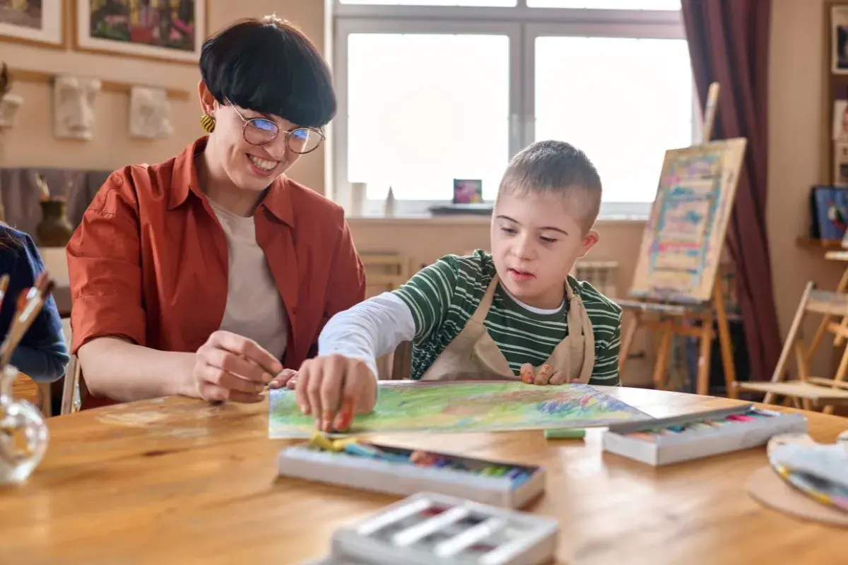Lehrerin unterstützt Kind mit Down-Syndrom beim Malen. Ein Beispiel für Förderung bei Autismus bei Kleinkindern.