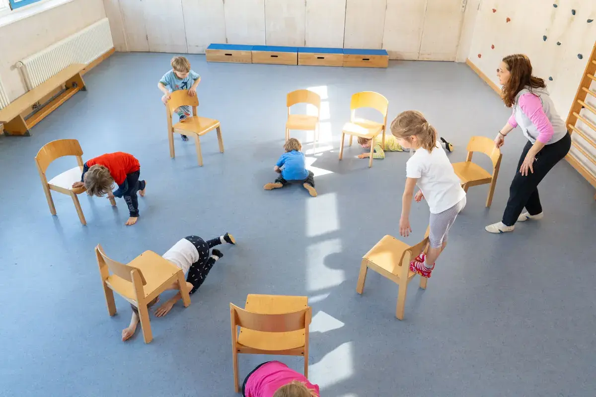 Kinder spielen in der Turnhalle, nutzen Stühle für kreative Ideen und Bewegung.
