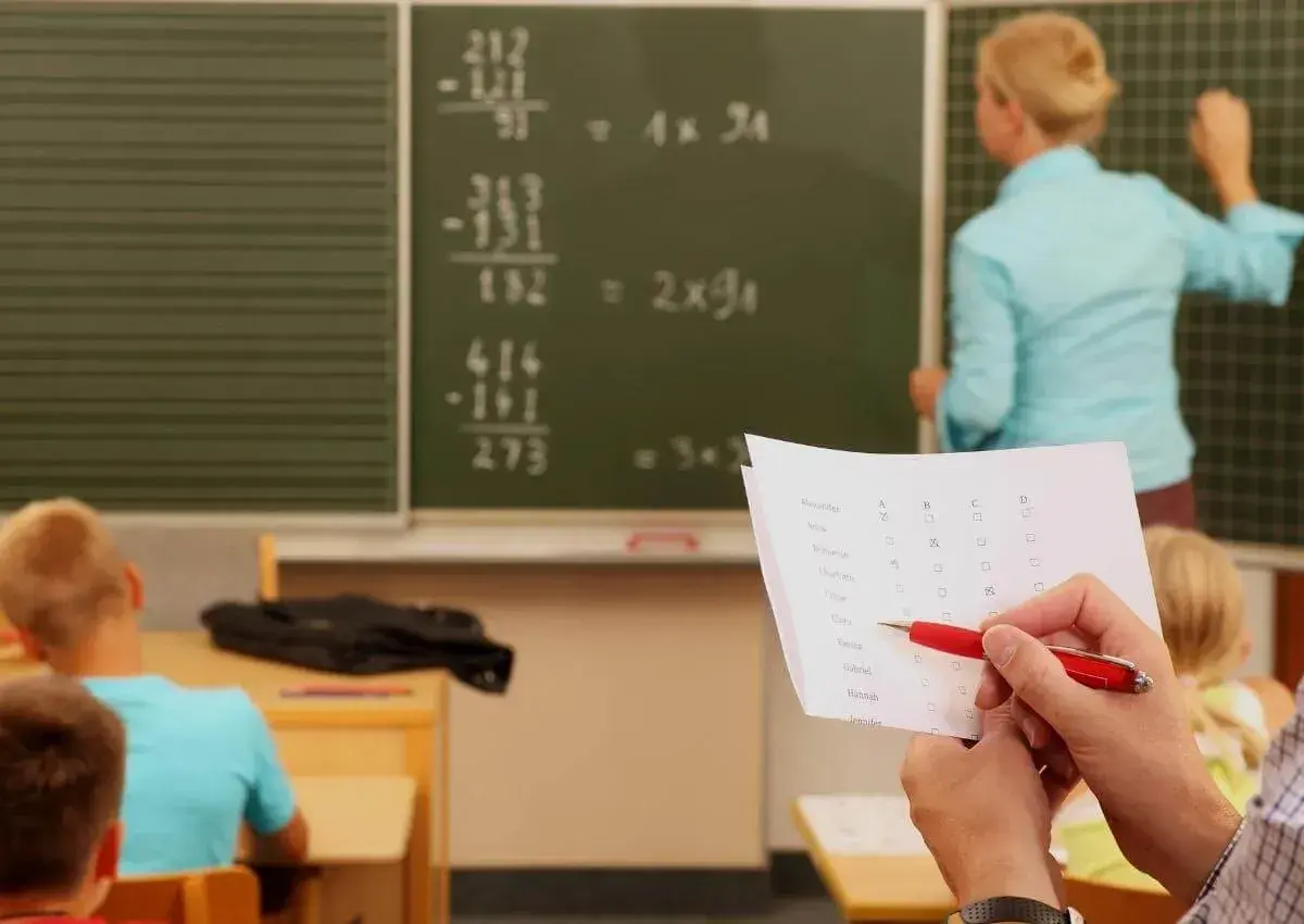 Lehrerin erklärt Mathe an Tafel. Ein Blatt mit Namen und Kästchen liegt bereit, wie bei dienstliche Beurteilung Lehrer Beispiele.