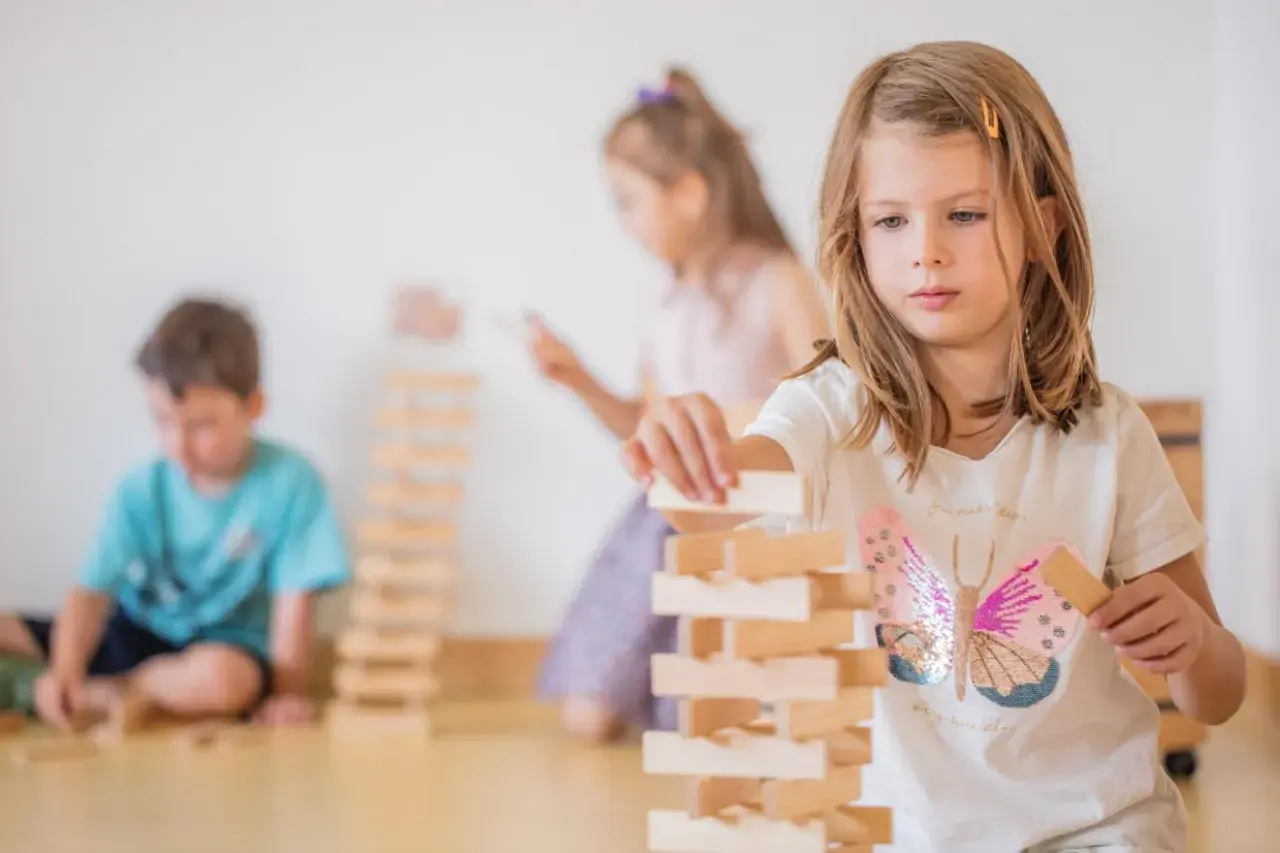 Kinder gestalten eine Bauecke im Kindergarten. Ein Mädchen baut konzentriert einen Turm aus Holzklötzen.