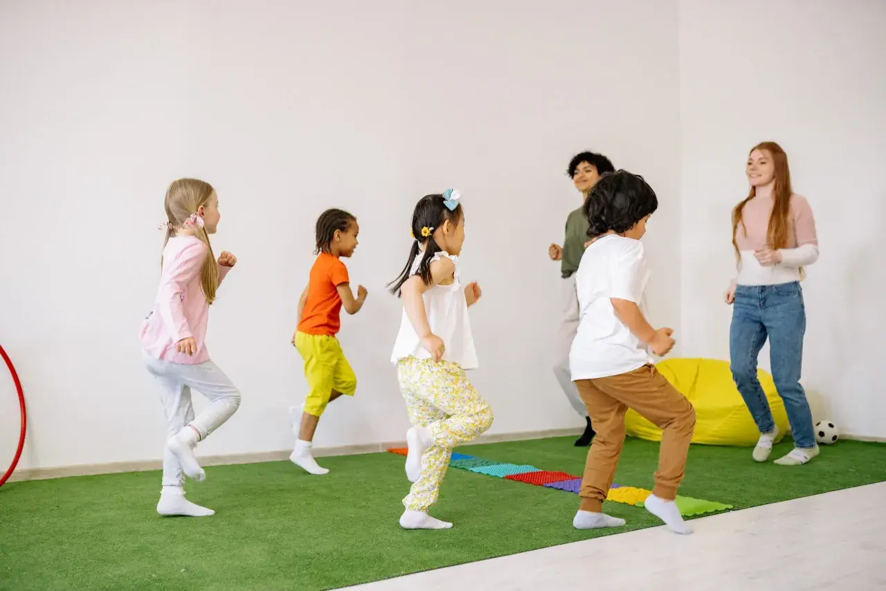 Kindergartenkinder rennen fröhlich über den grünen Teppich, als ob sie Tiere wären. Die Erzieherinnen machen mit bei den lustigen Bewegungsspielen.