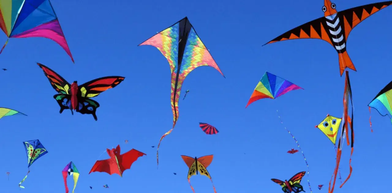 Bunte Drachen in Form von Schmetterlingen, Fledermäusen und geometrischen Mustern steigen im blauen Himmel auf. Ein Fest des Drachenbastelns!