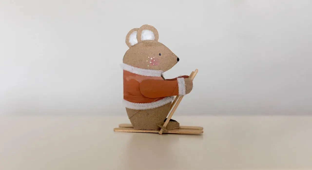 Süße Maus beim Skifahren, ein tolles Beispiel für Basteln mit Papprollen. Sie trägt einen orangen Pullover und hält Skistöcke.