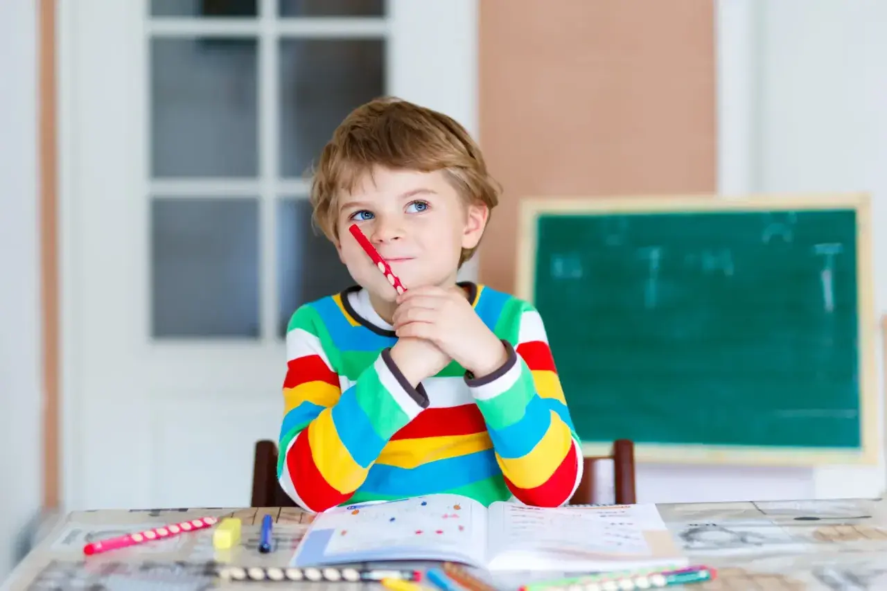 Ein Junge mit einem roten Stift im Mund denkt nach. Seine Kleidung und die Lernmaterialien deuten auf eine mögliche **Hochbegabung bei Kindern** hin.