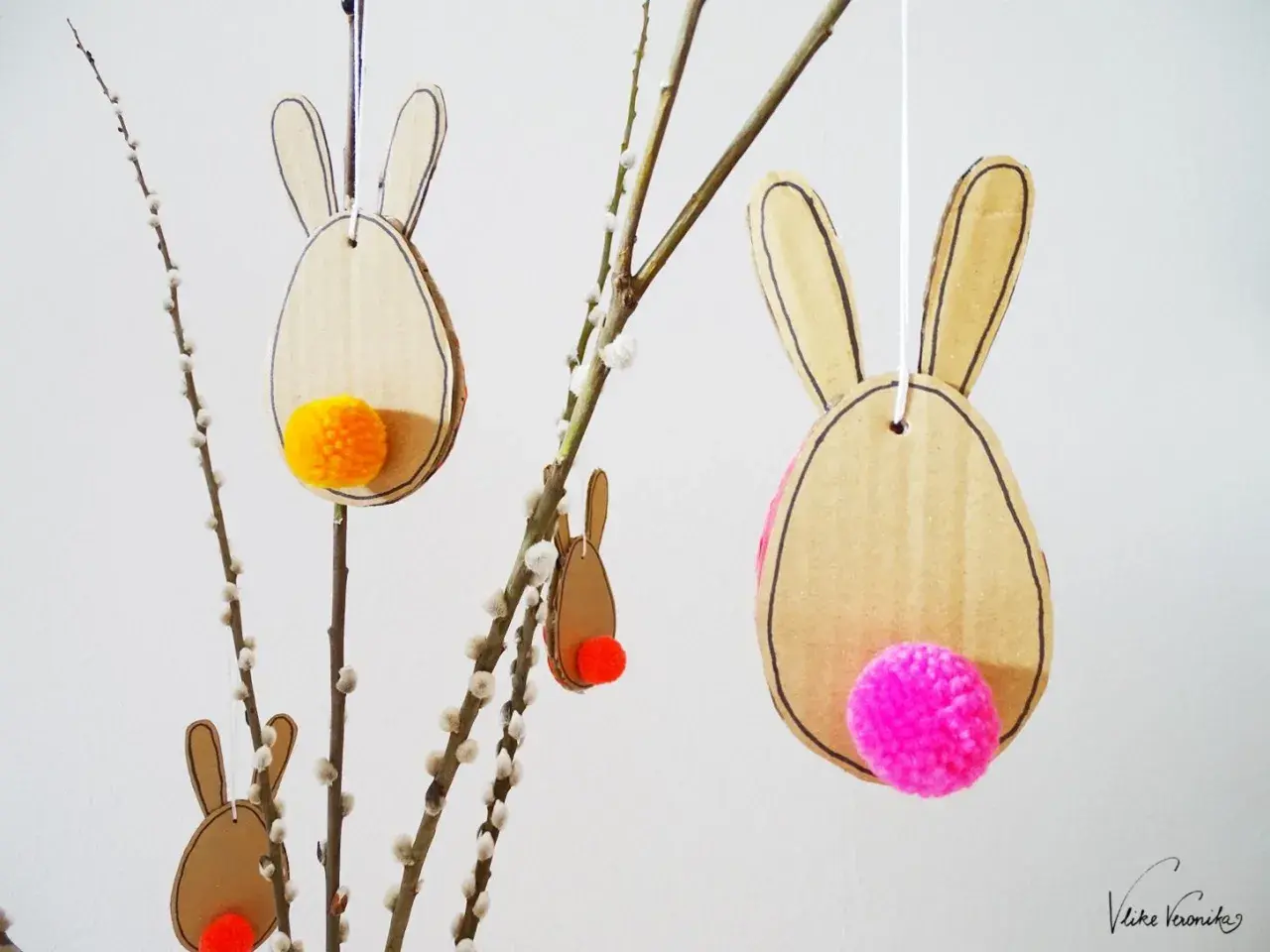 Osterhasen aus Pappe mit bunten Bommeln als Schwänzen, perfekt zum Basteln Ostern Kinder. Hängen an Weidenkätzchen-Zweigen.