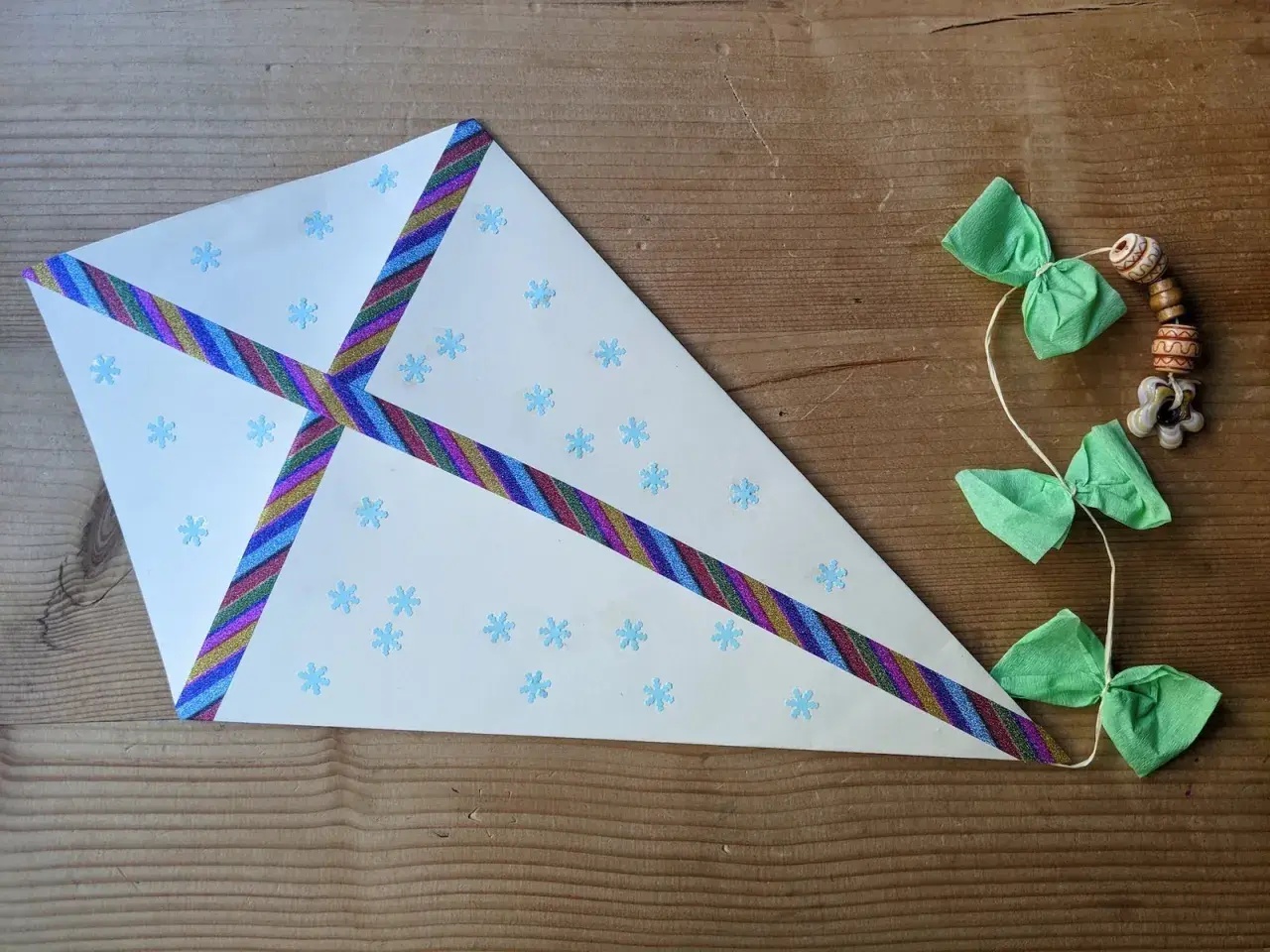 Ein selbstgebastelter Drachen aus Tonpapier mit Schneeflocken und bunten Bändern. Die Schnur hat kleine grüne Schleifen und Holzperlen.