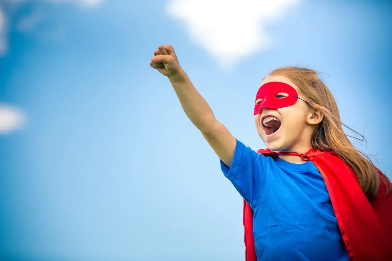 Ein kleines Mädchen im Superheldenkostüm mit rotem Cape und Maske, bereit für den **ersten Schultag**.