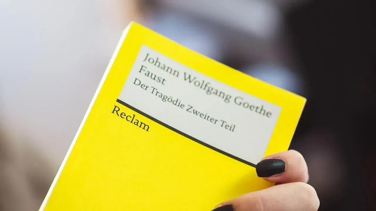 Ein gelbes Buch von Reclam mit Goethes Faust, Teil 2. Eine Hand mit schwarzen Nägeln hält es. Textanalyse Deutsch.